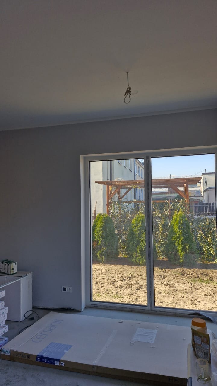 Wnętrze pokoju z świeżo pomalowanymi szarymi ścianami, widok na ogród przez duże okno, na podłodze leżą opakowania płytek ceramicznych marki Cersanit.