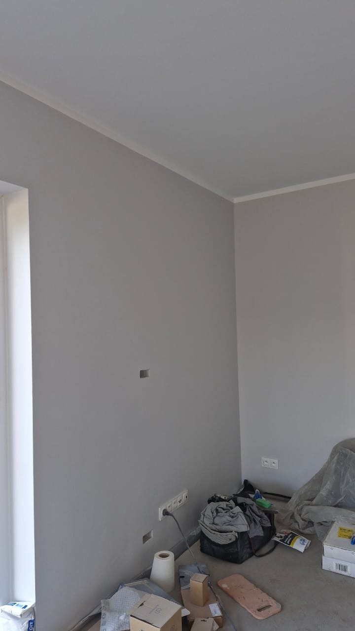 Pokój po malowaniu na szaro, z widocznymi gniazdkami elektrycznymi, puszką instalacyjną, listwą przypodłogową i drobnymi pozostałościami po pracach wykończeniowych na podłodze.