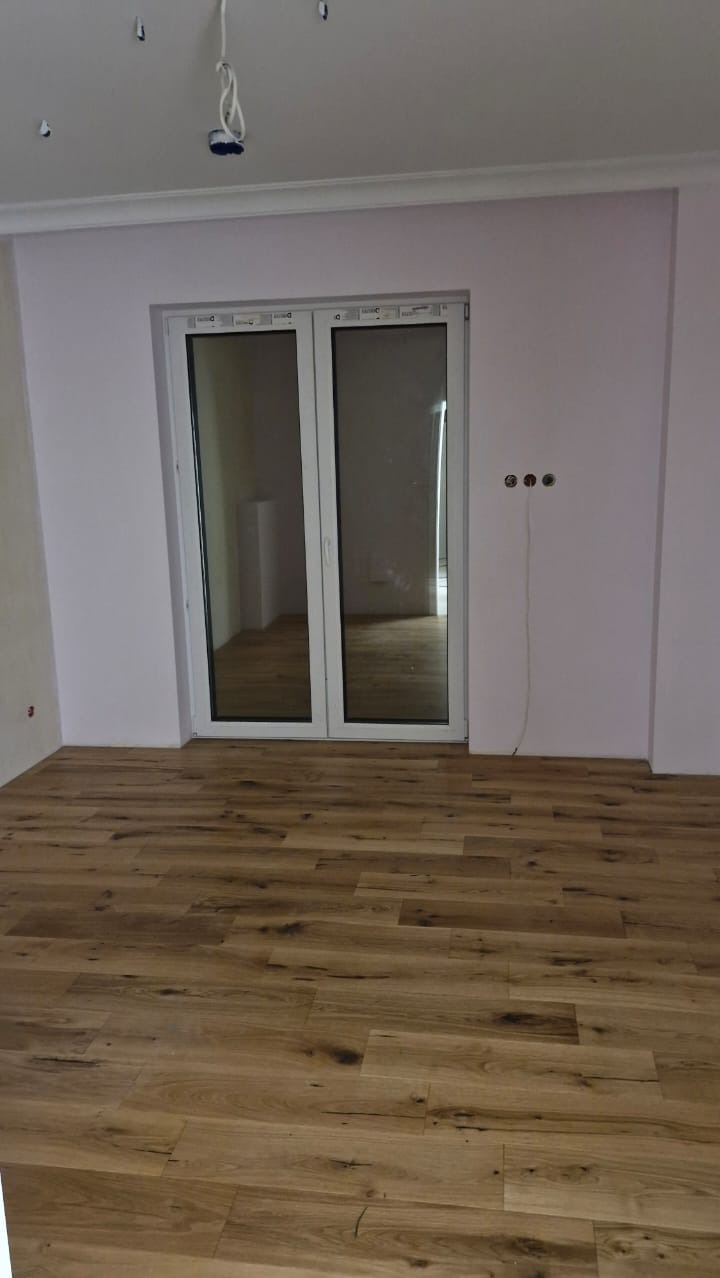 Ułożone panele podłogowe w pokoju z białymi drzwiami balkonowymi i przygotowanymi punktami elektrycznymi na ścianie.