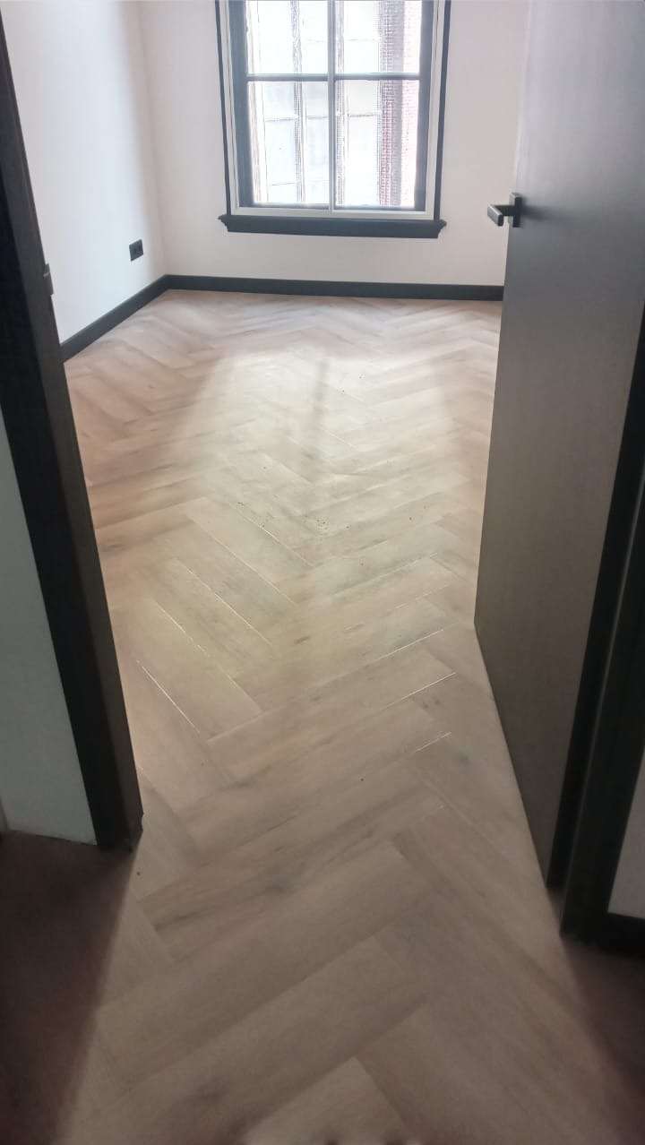 Jasna, drewniana podłoga ułożona w jodełkę w pustym pokoju z oknem i ciemnymi listwami przypodłogowymi, widziana z perspektywy otwartych drzwi.