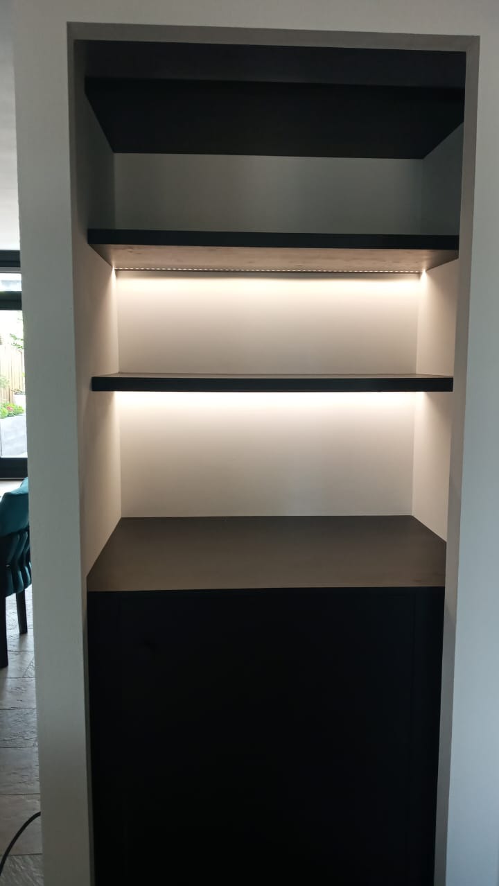 Wnęka w ścianie z czarnymi półkami podświetlonymi ledowym światłem, widok z boku na fragment salonu z turkusowym krzesłem.