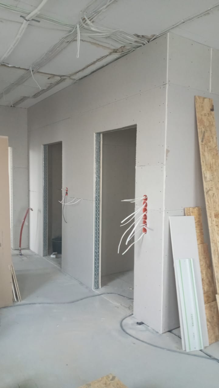 Wnętrze w trakcie prac wykończeniowych, widoczne ściany z płyt kartonowo-gipsowych z metalowymi profilami, instalacja elektryczna w peszlach, płyty OSB oparte o ścianę i surowy beton na podłodze.