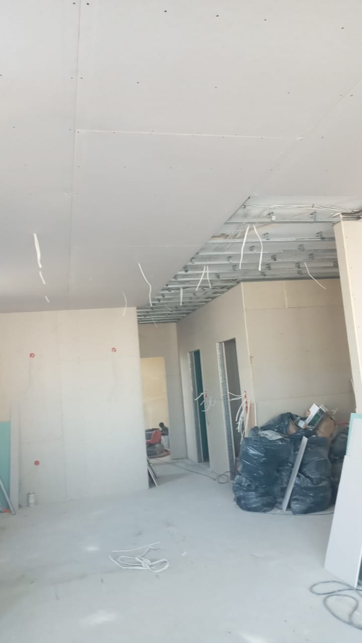 Wnętrze w trakcie montażu płyt kartonowo-gipsowych: widoczne stelaże sufitowe, częściowo zamontowane płyty, kable elektryczne zwisające z sufitu, worki z odpadami budowlanymi i płyty gipsowe oparte...