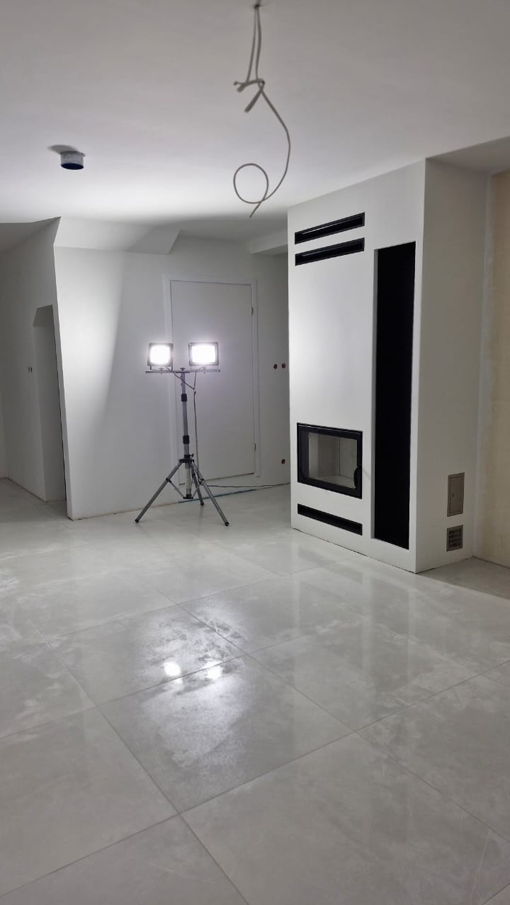 Wnętrze w trakcie prac wykończeniowych: widoczna lampa budowlana na trójnogu, fragment ściany z obudową kominka w kolorze białym z czarnymi elementami dekoracyjnymi oraz przewody elektryczne...