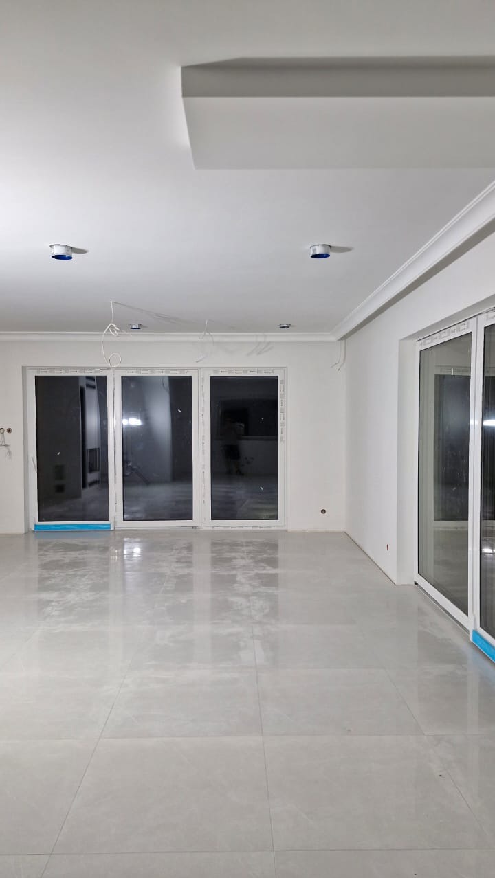 Jasne, przestronne wnętrze z dużymi oknami tarasowymi, wykończone płytkami podłogowymi w odcieniach szarości, widoczne przygotowania do montażu oświetlenia sufitowego oraz listwy dekoracyjne...