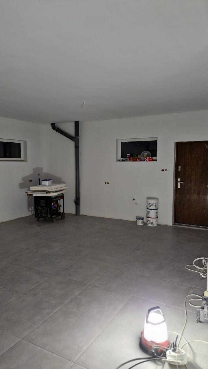 Wnętrze pokoju w trakcie odświeżania: widoczne świeżo pomalowane ściany na biało, szare płytki na podłodze, oraz przygotowane farby i agregat malarski.