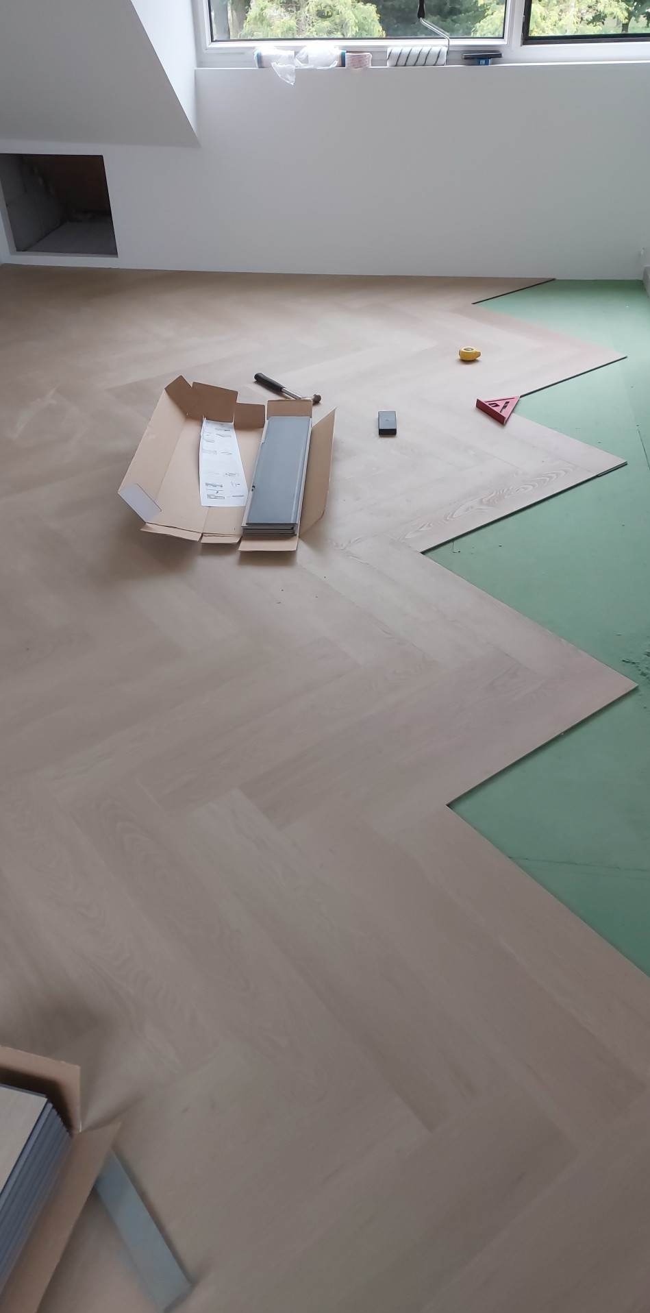 Układanie jasnych paneli podłogowych w jodełkę na zielonej podkładce, widoczne narzędzia i otwarte opakowanie paneli.