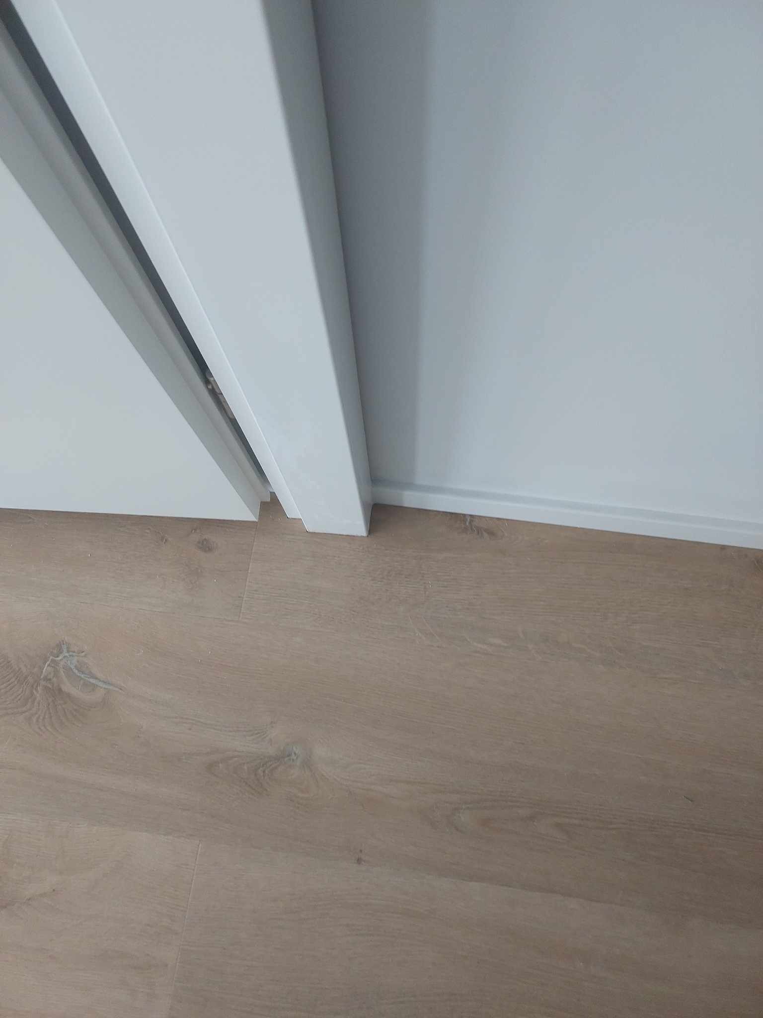 Fragment wnętrza z jasnymi panelami podłogowymi, białą listwą przypodłogową i fragmentem białych drzwi przesuwnych.