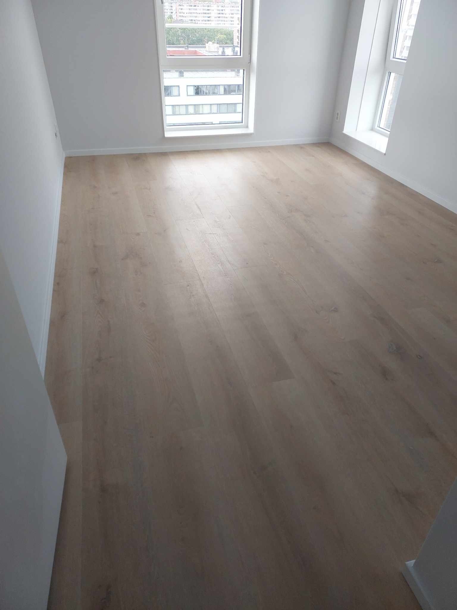 Jasne panele podłogowe w pustym, nowym pokoju z dwoma oknami, widok na miasto za oknem, minimalistyczny design.