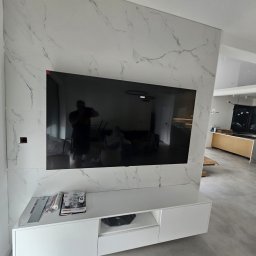 Michal Halas - Wykończona ściana z płyt imitujących marmur, z zawieszonym telewizorem i białą szafką RTV. Widoczne narzędzia i materiały budowlane.