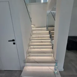 Nowoczesne schody z białego marmuru z podświetleniem LED, szklane balustrady, minimalistyczny design. Butelka wody na pierwszym stopniu.