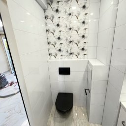 Michal Halas - Wykończona toaleta w bieli z akcentem kwiatowym nad zabudową. Czarna armatura i deska sedesowa. Podłoga wyłożona płytkami imitującymi marmur.