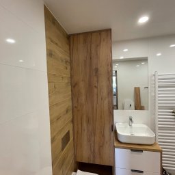 Michal Halas - Nowoczesna łazienka po remoncie: białe płytki, drewnopodobne panele, szafka z umywalką i toaleta. Minimalistyczny design, ciepłe oświetlenie.