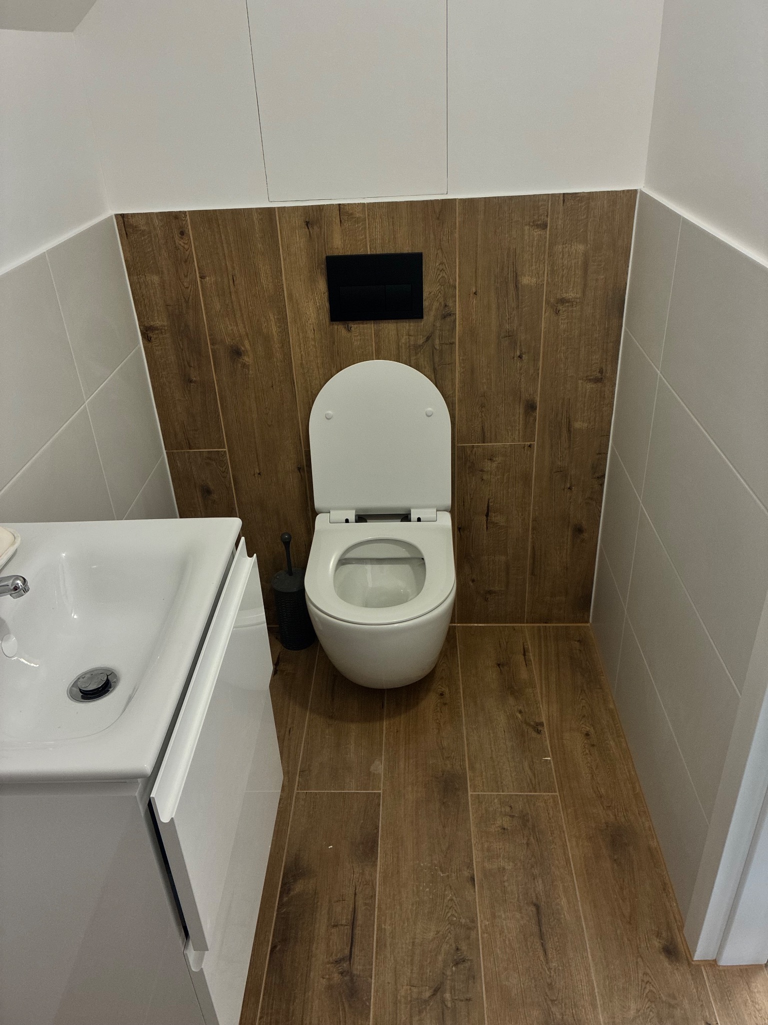 Nowoczesna łazienka po remoncie: biała umywalka z szafką, toaleta z deską, drewnopodobne płytki na podłodze i ścianie, szare płytki na ścianach.