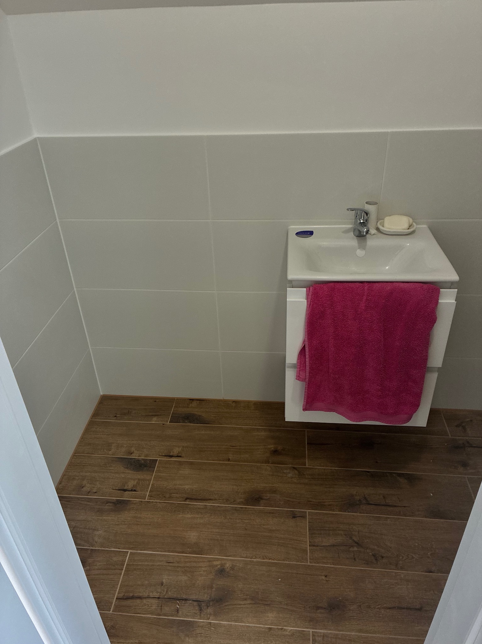 Jasna łazienka po remoncie: drewnopodobne płytki na podłodze, białe płytki na ścianach, umywalka z szafką i różowy ręcznik. Minimalistyczny design.