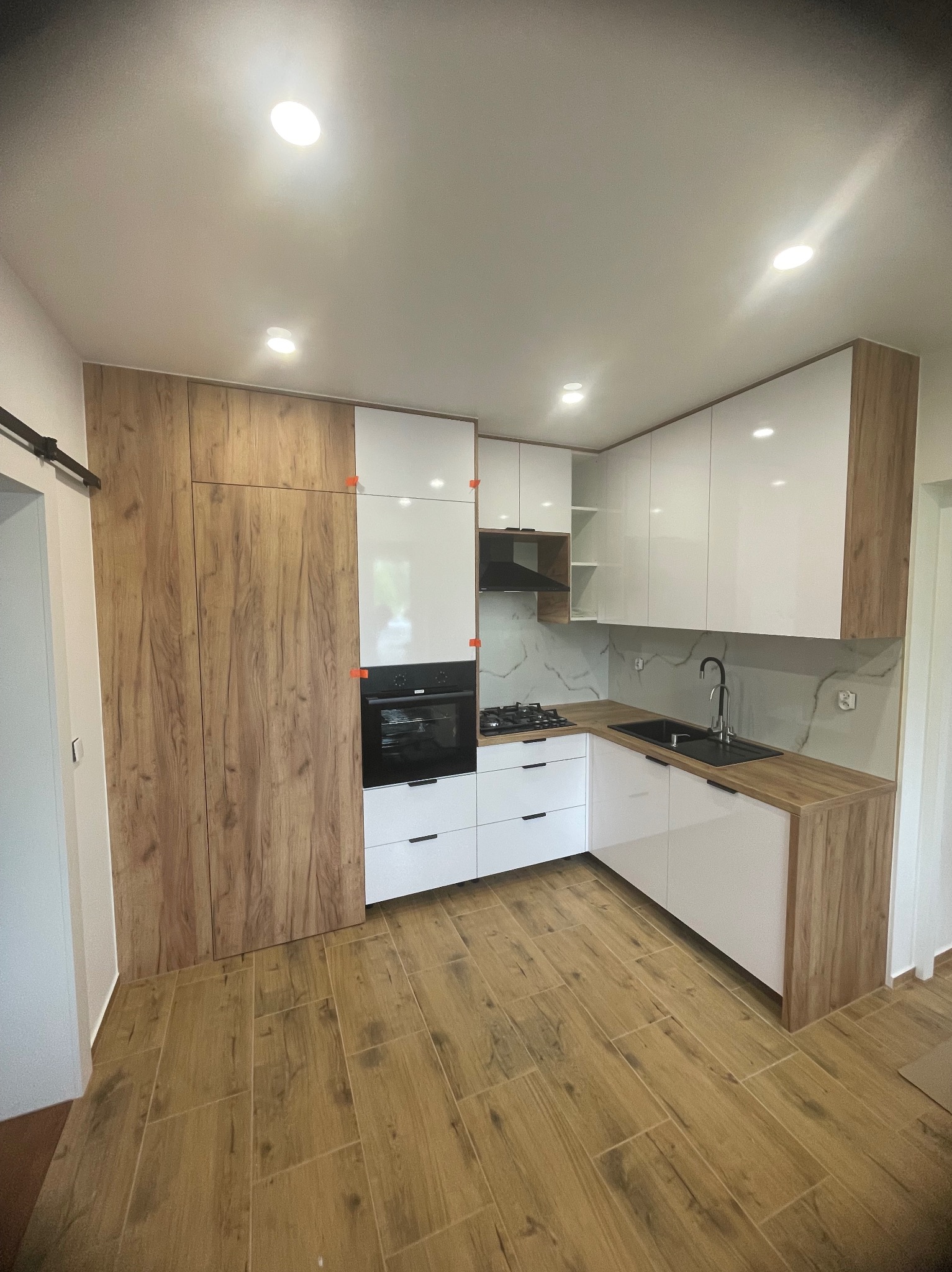 Nowoczesna kuchnia w Nowym Sączu: białe szafki z drewnianymi akcentami, czarny zlew i sprzęt AGD. Jasne płytki podłogowe imitujące drewno.