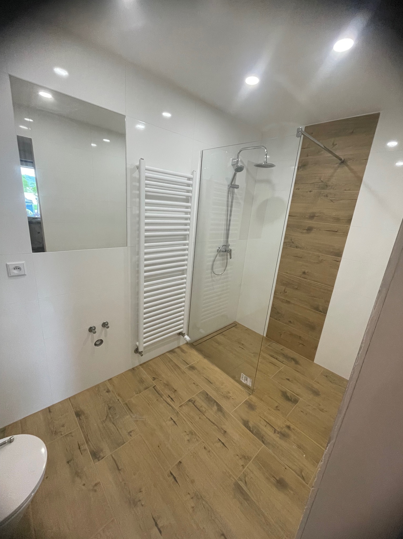 Nowoczesna łazienka po remoncie: kabina prysznicowa ze szklaną ścianą, grzejnik drabinkowy, lustro i płytki imitujące drewno na podłodze. Białe płytki na ścianach.
