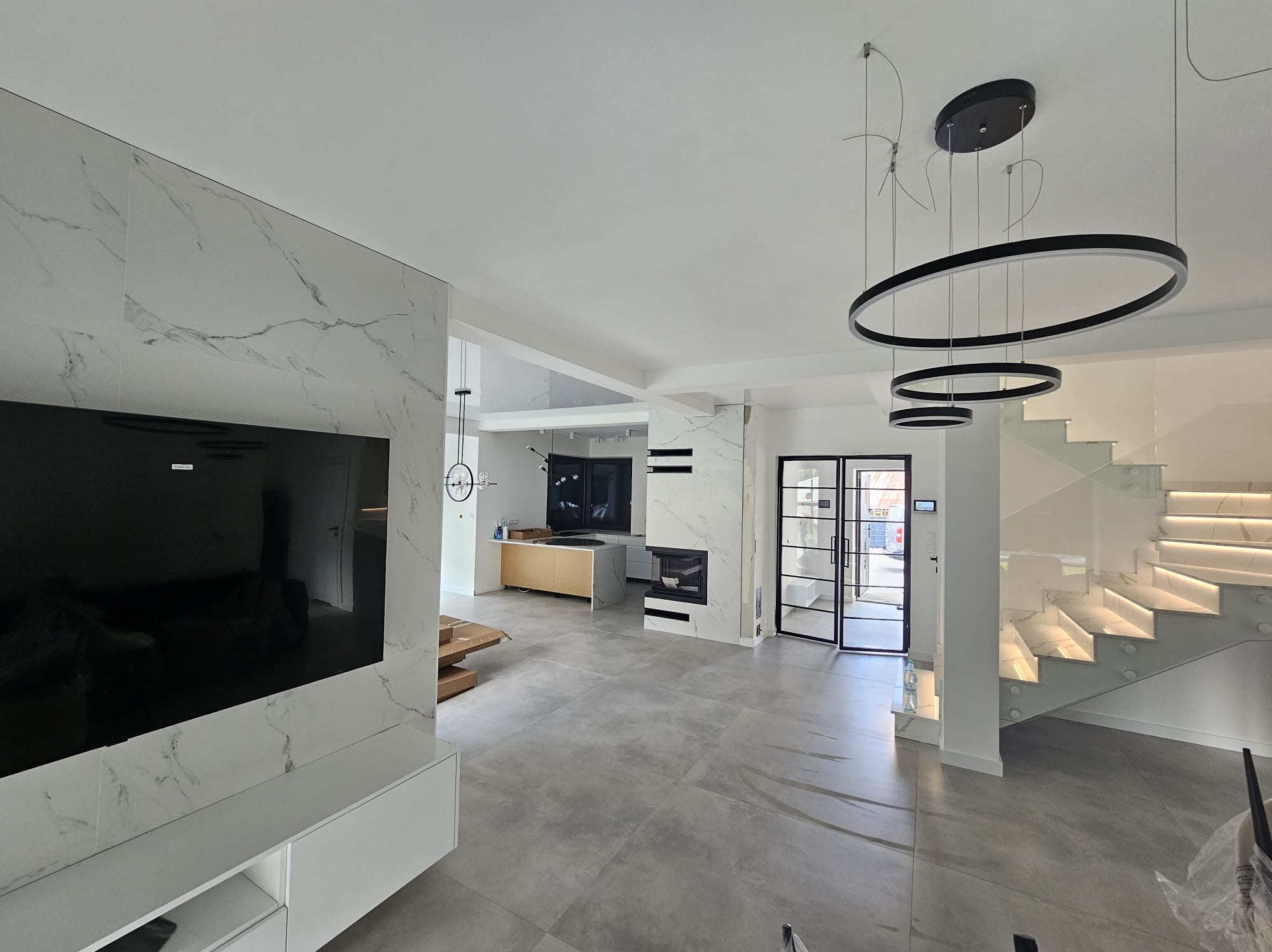 Nowoczesne wnętrze z marmurową ścianą TV, szarymi płytkami podłogowymi i schodami z podświetleniem LED. Widoczny kominek i okrągłe lampy wiszące.