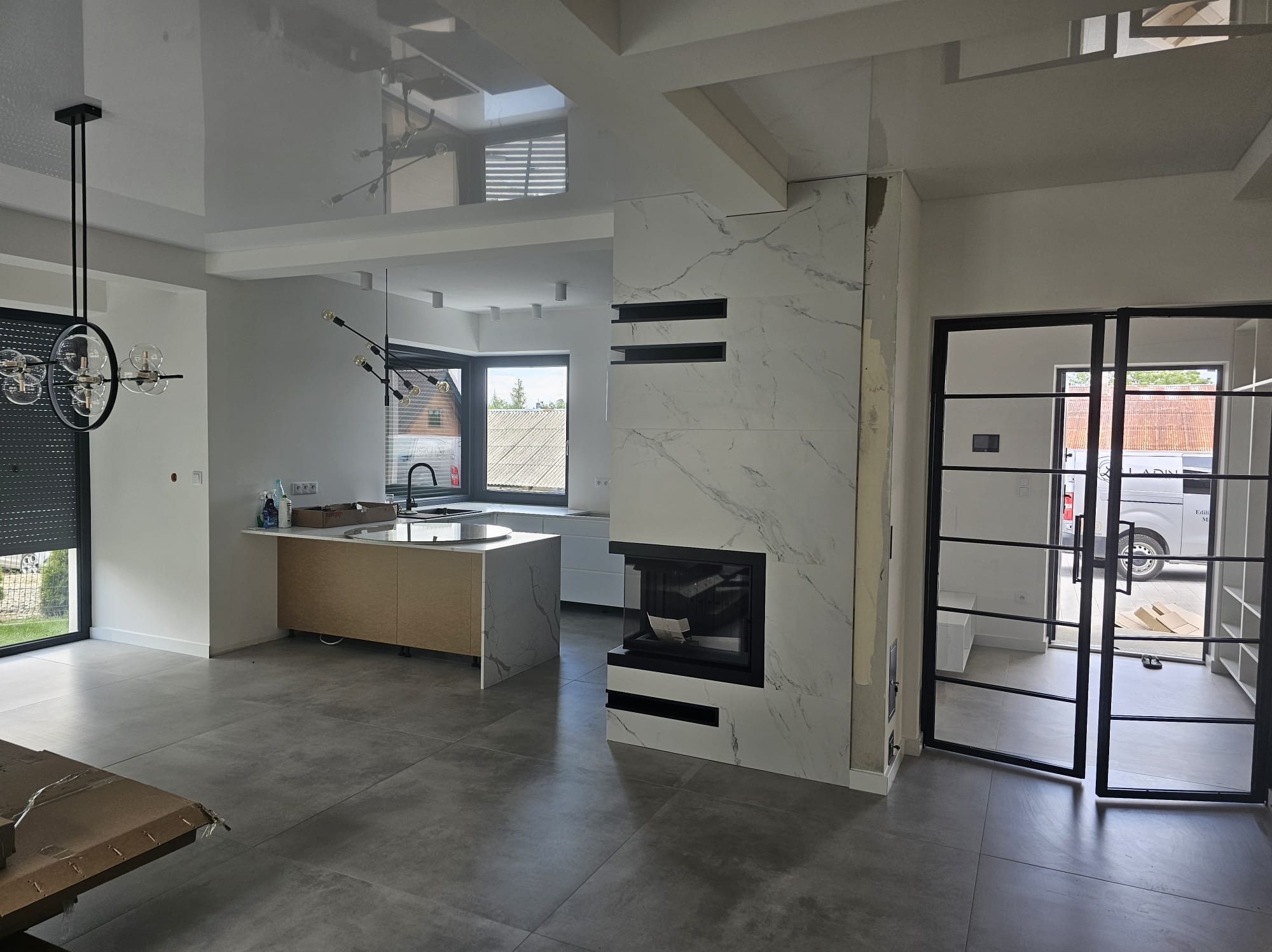 Nowoczesne wnętrze z marmurowym kominkiem, wyspą kuchenną i szklanymi drzwiami. Szara podłoga i białe ściany tworzą minimalistyczny design. Widoczne elementy oświetlenia.