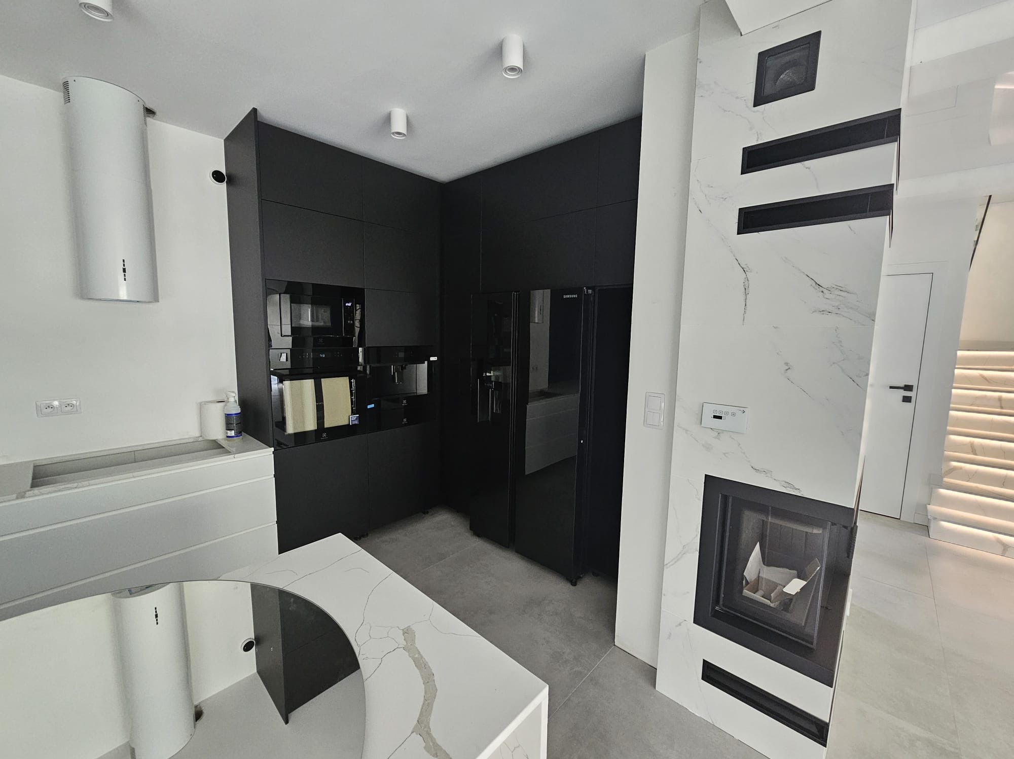 Nowoczesna kuchnia z czarnymi szafkami i sprzętami, biały blat z marmurowym wzorem, kominek obudowany płytami imitującymi marmur, schody z podświetleniem.