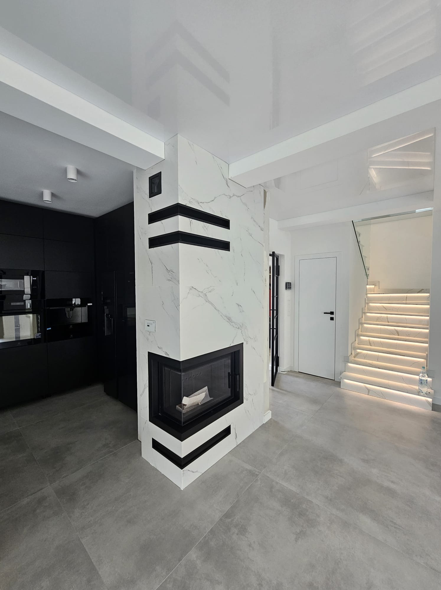 Nowoczesne wnętrze z marmurowym kominkiem i podświetlanymi schodami. Minimalistyczna kuchnia w czerni, szare płytki podłogowe i białe ściany tworzą elegancką przestrzeń.