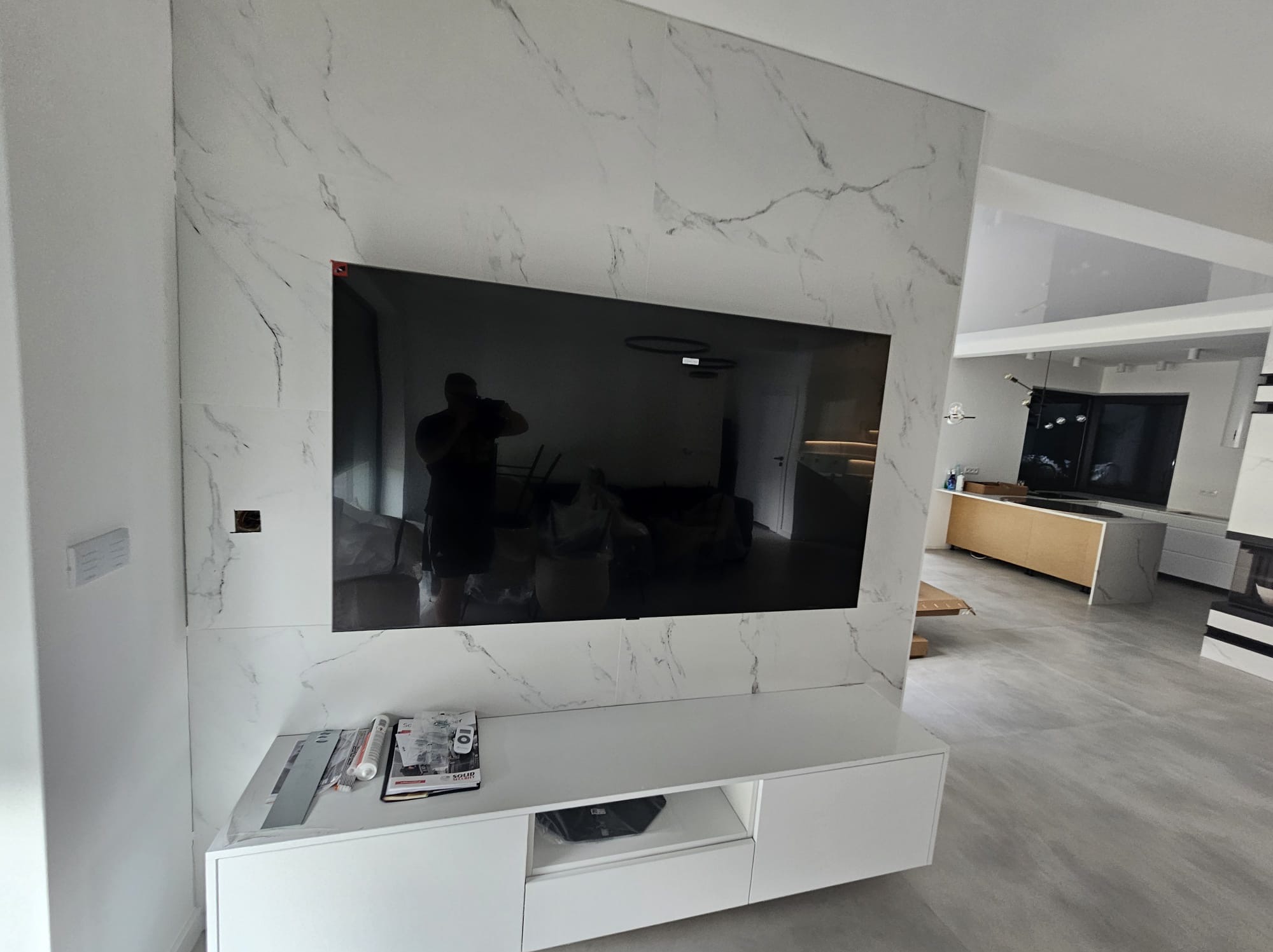 Wykończona ściana z płyt imitujących marmur, z zawieszonym telewizorem i białą szafką RTV. Widoczne narzędzia i materiały budowlane.
