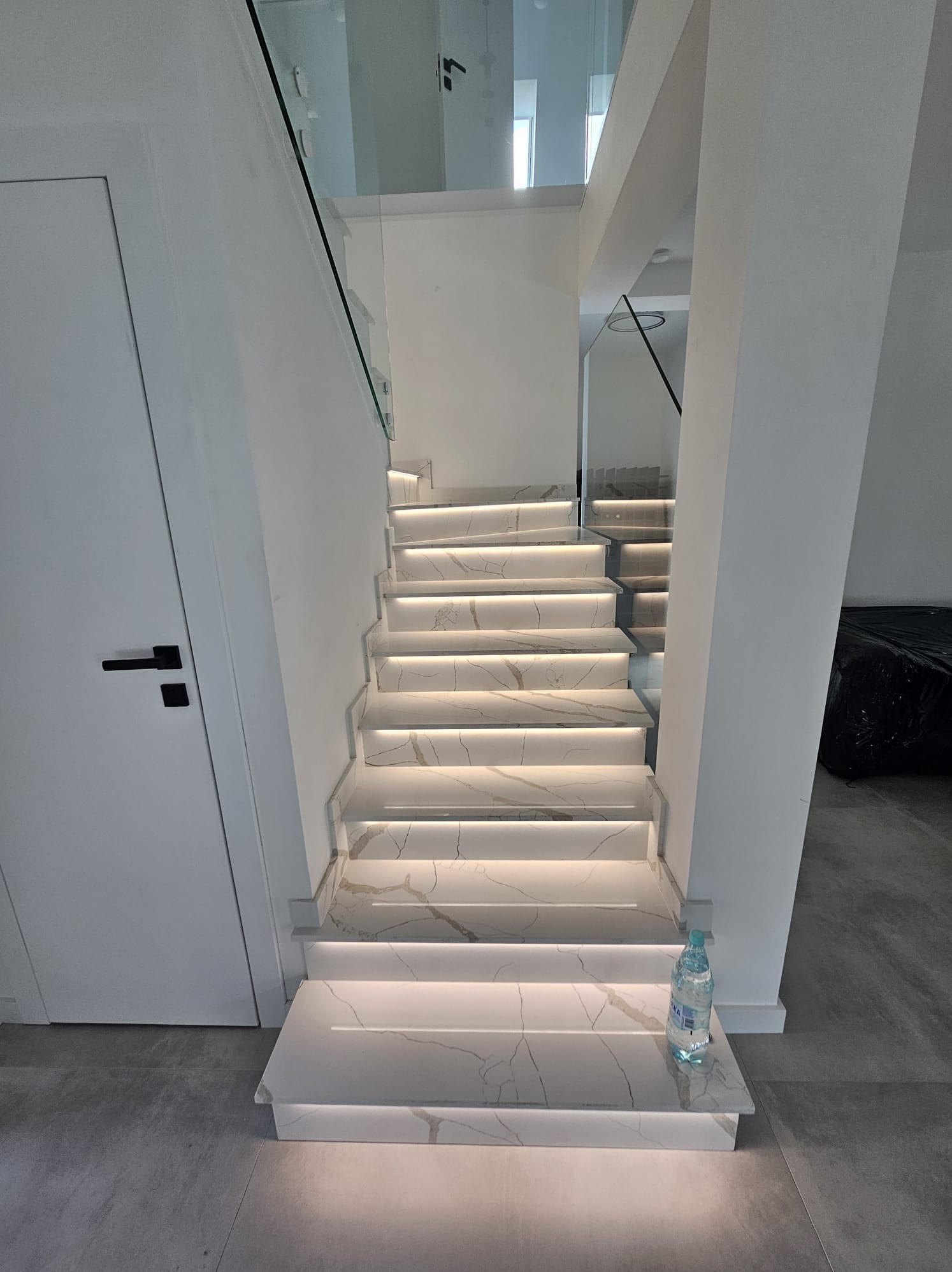 Nowoczesne schody z białego marmuru z podświetleniem LED, szklane balustrady, minimalistyczny design. Butelka wody na pierwszym stopniu.