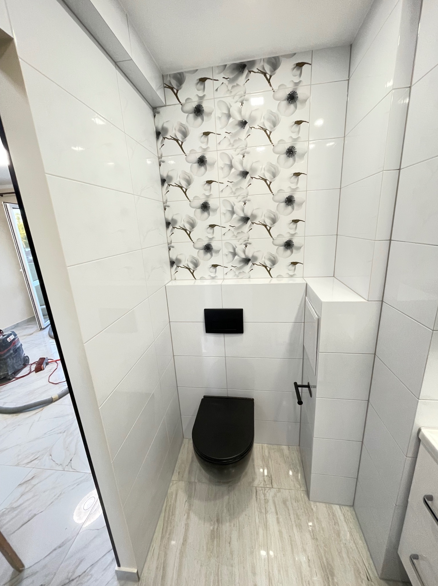 Wykończona toaleta w bieli z akcentem kwiatowym nad zabudową. Czarna armatura i deska sedesowa. Podłoga wyłożona płytkami imitującymi marmur.