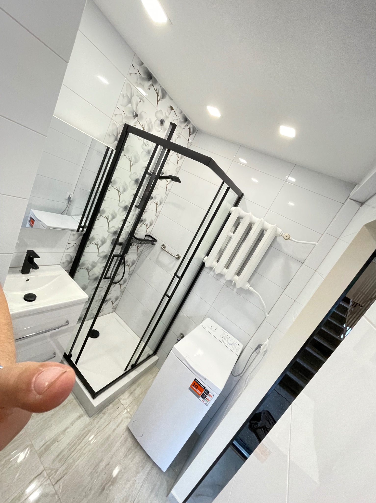 Nowoczesna łazienka po remoncie: białe płytki, kabina prysznicowa z czarnymi ramami, umywalka z szafką i pralka. Na ścianie płytki z motywem kwiatowym.