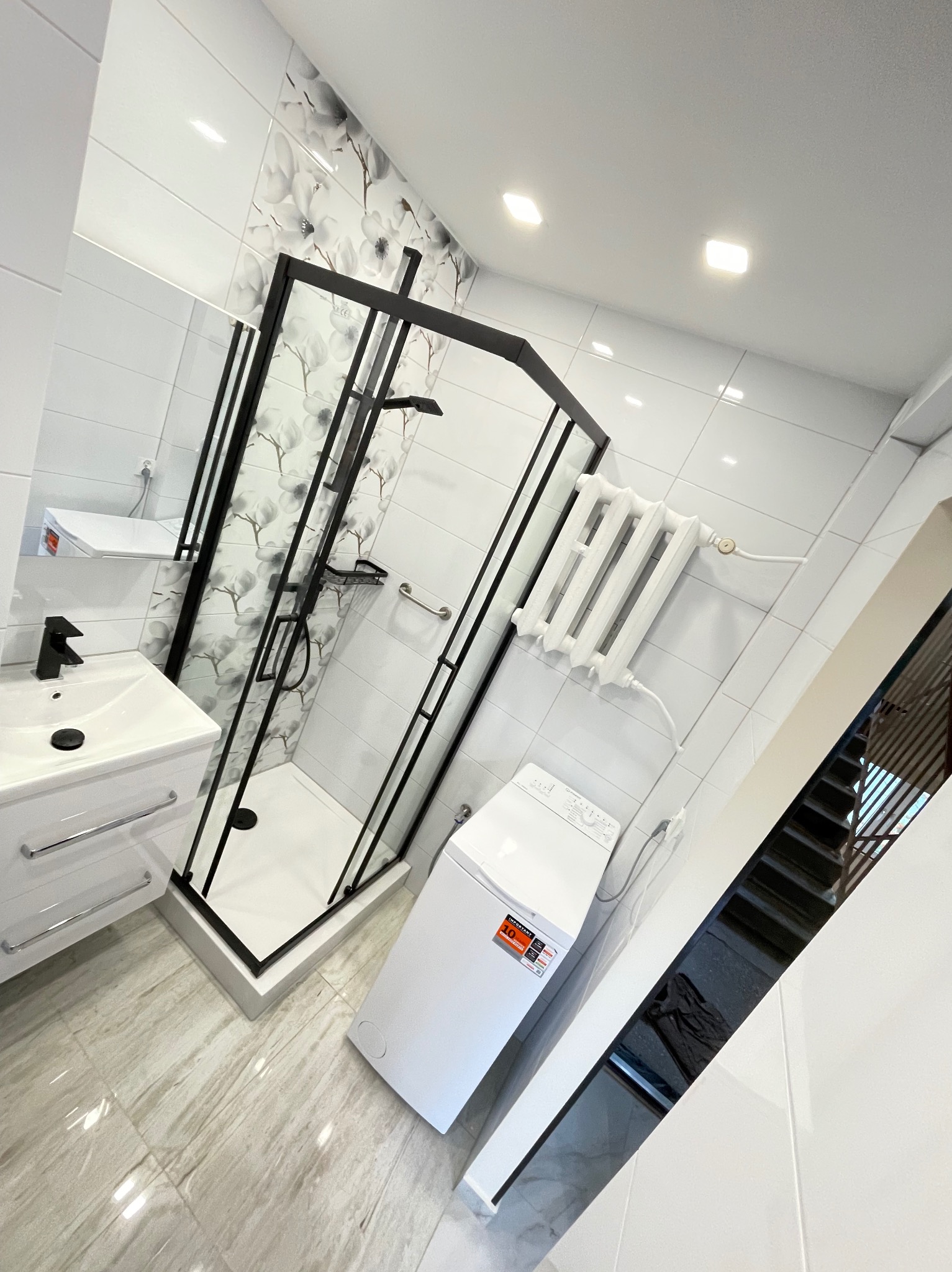 Nowoczesna łazienka po remoncie: białe płytki, kabina prysznicowa z czarną ramą, umywalka z szafką, pralka i grzejnik. Kwiatowy motyw na ścianie dodaje elegancji.