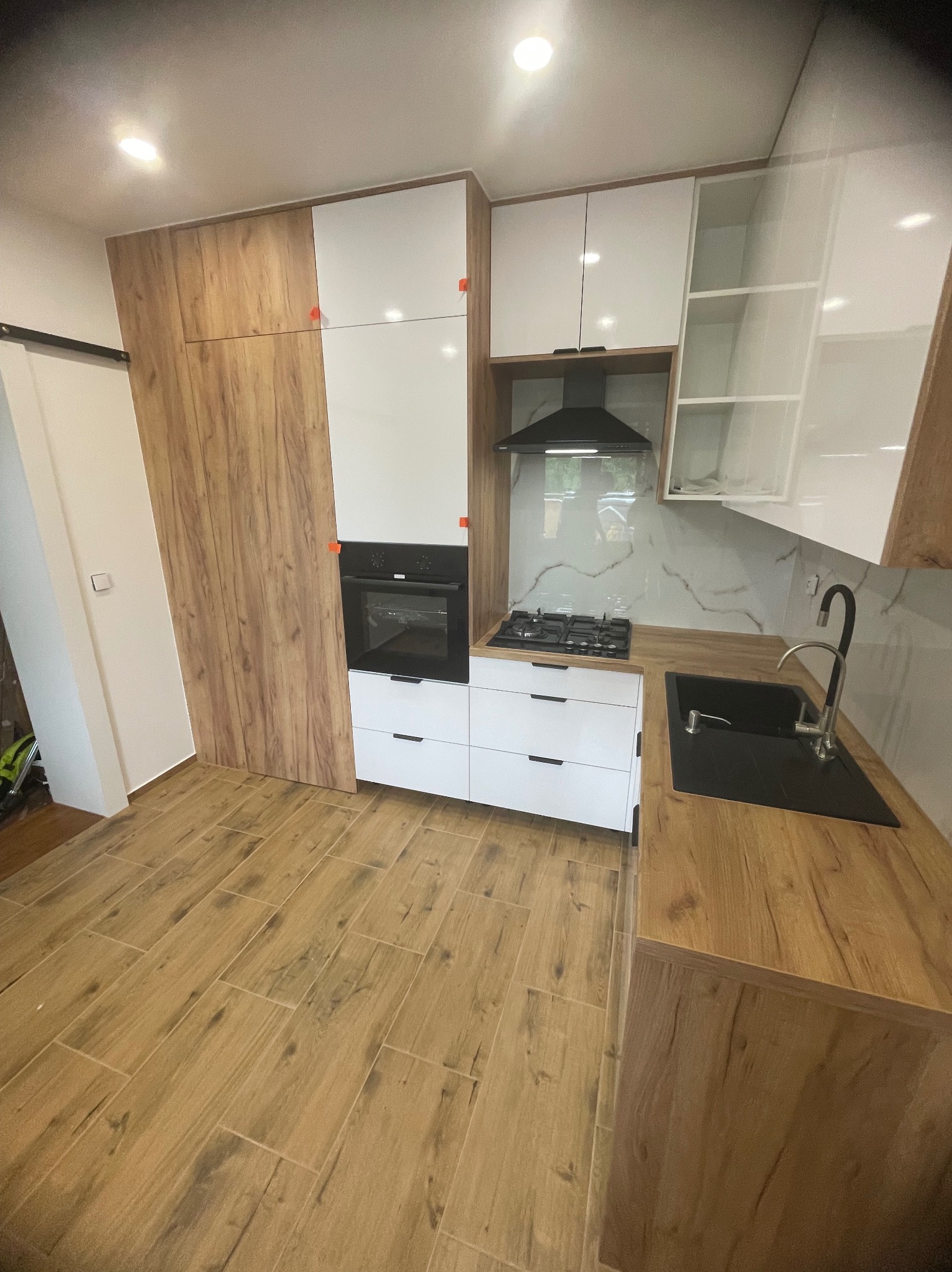 Nowoczesna kuchnia z drewnianymi i białymi szafkami, czarnym zlewem i płytą gazową. Podłoga wyłożona panelami imitującymi drewno. Widok na marmurowy backsplash.