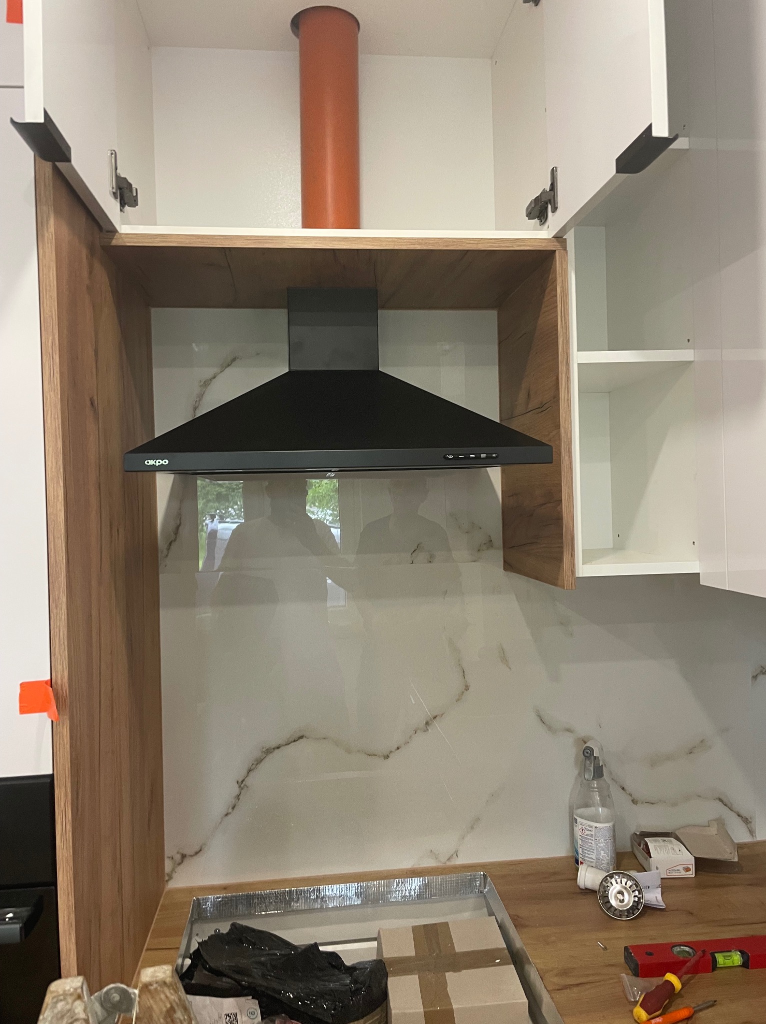 Czarny okap kuchenny marki Akpo zamontowany nad blatem z marmurową płytą. Widoczne otwarte szafki z białymi frontami i drewnianymi bokami. Pomarańczowa rura wentylacyjna.