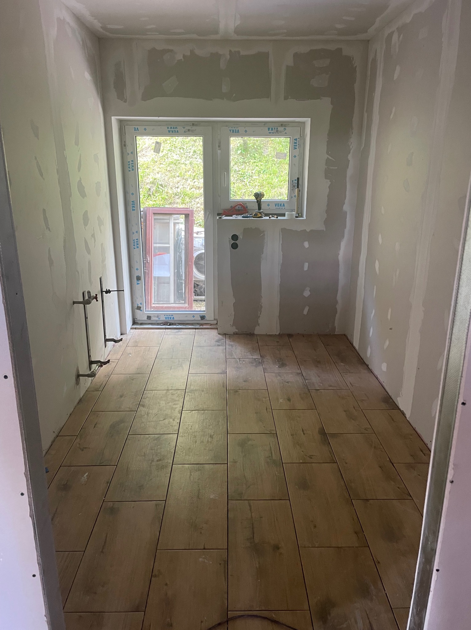 Wnętrze w trakcie remontu: ułożona podłoga z płytek imitujących drewno, ściany przygotowane pod malowanie, widoczne instalacje hydrauliczne i okno z widokiem na zieleń.