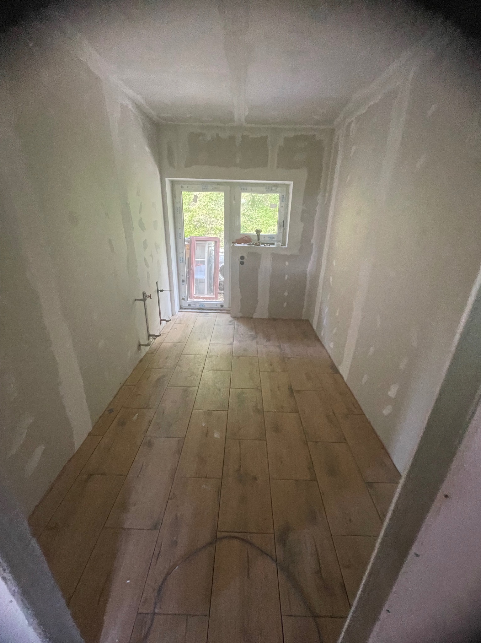 Wnętrze w trakcie remontu: ułożona podłoga z płytek imitujących drewno, ściany przygotowane pod malowanie, widoczne instalacje. Okno z widokiem na zieleń.