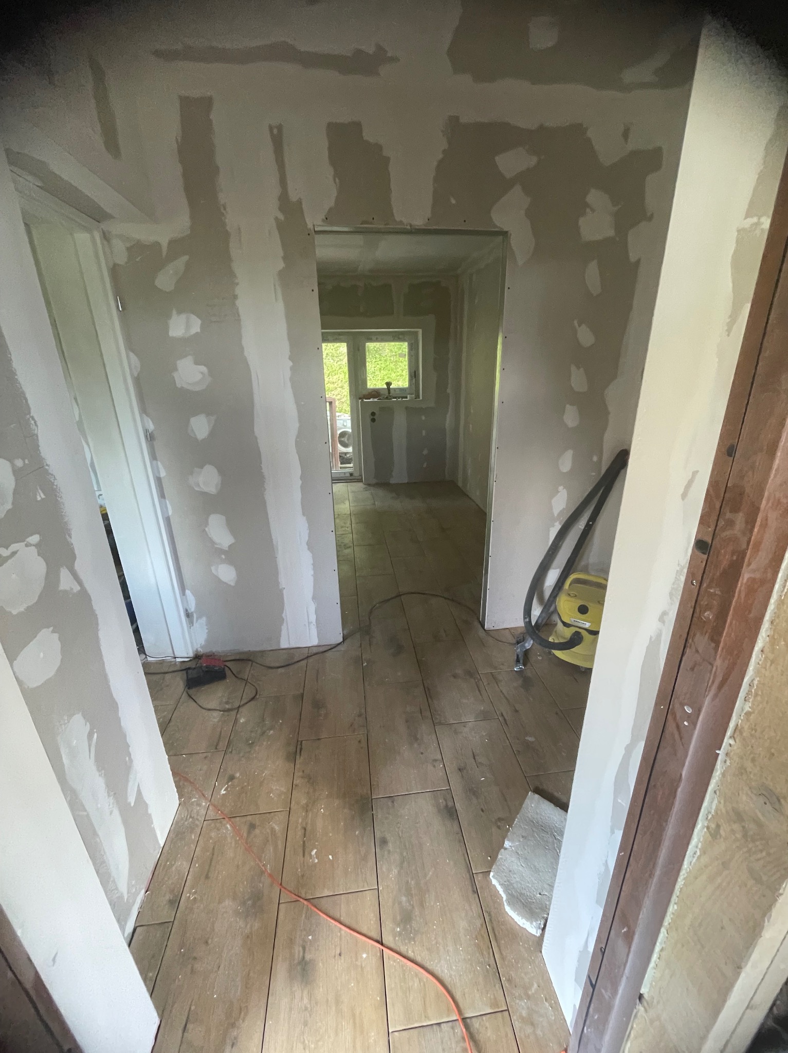 Wnętrze w trakcie prac wykończeniowych: ściany z płyt karton-gips, widoczne łączenia, drewnopodobna podłoga, narzędzia budowlane. Widok na okno w głębi.