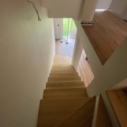 Aliaksei Shabaila - Drewniane schody w jasnym wnętrzu, widok z góry. Minimalistyczny design, białe ściany, naturalne światło wpadające przez okno. Częściowo widoczne stopy na dole kadru.