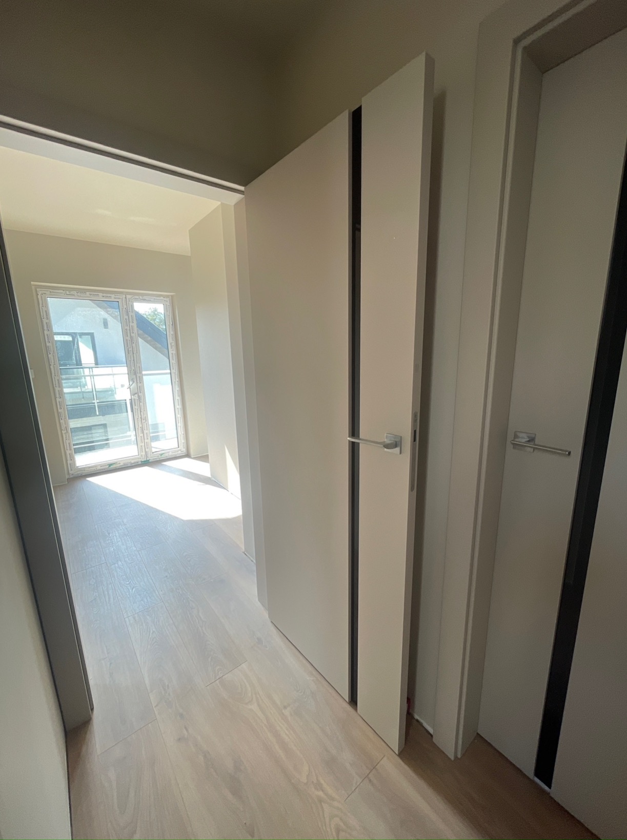 Wnętrze mieszkania z drewnianą podłogą, białymi drzwiami z czarnymi wstawkami i widokiem na balkon przez okno balkonowe.