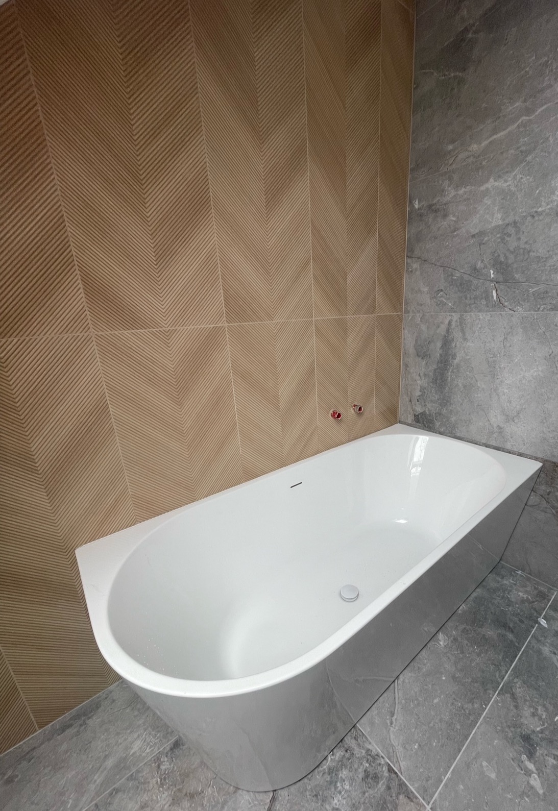 Biała wanna narożna na tle ściany wyłożonej płytkami w odcieniach beżu z wzorem chevron i szarymi płytkami imitującymi marmur.