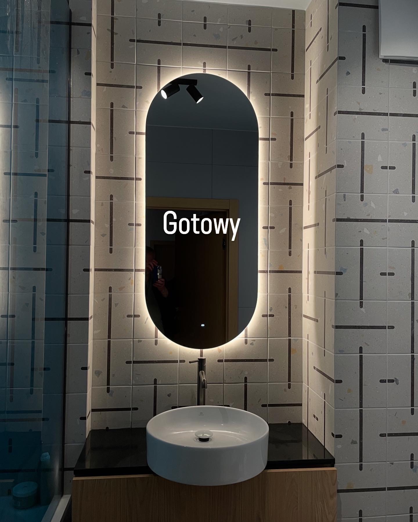 Nowoczesna łazienka z owalnym lustrem LED, białą umywalką nablatową i płytkami w geometryczny wzór. Minimalistyczny design, jasne oświetlenie.