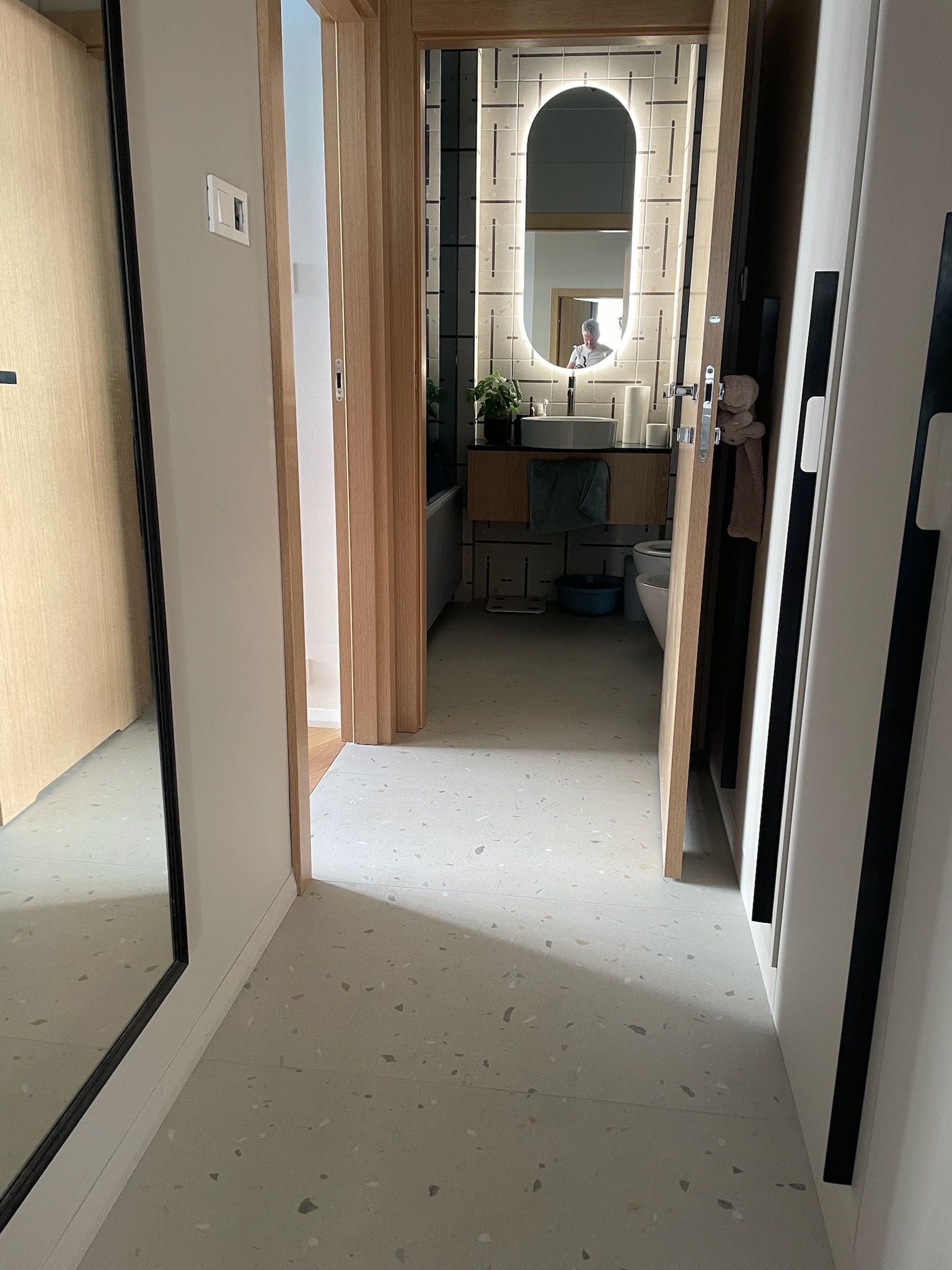 Wnętrze łazienki z podświetlanym lustrem owalnym, widok z korytarza. Jasna kolorystyka, minimalistyczny styl. Na podłodze lastryko.