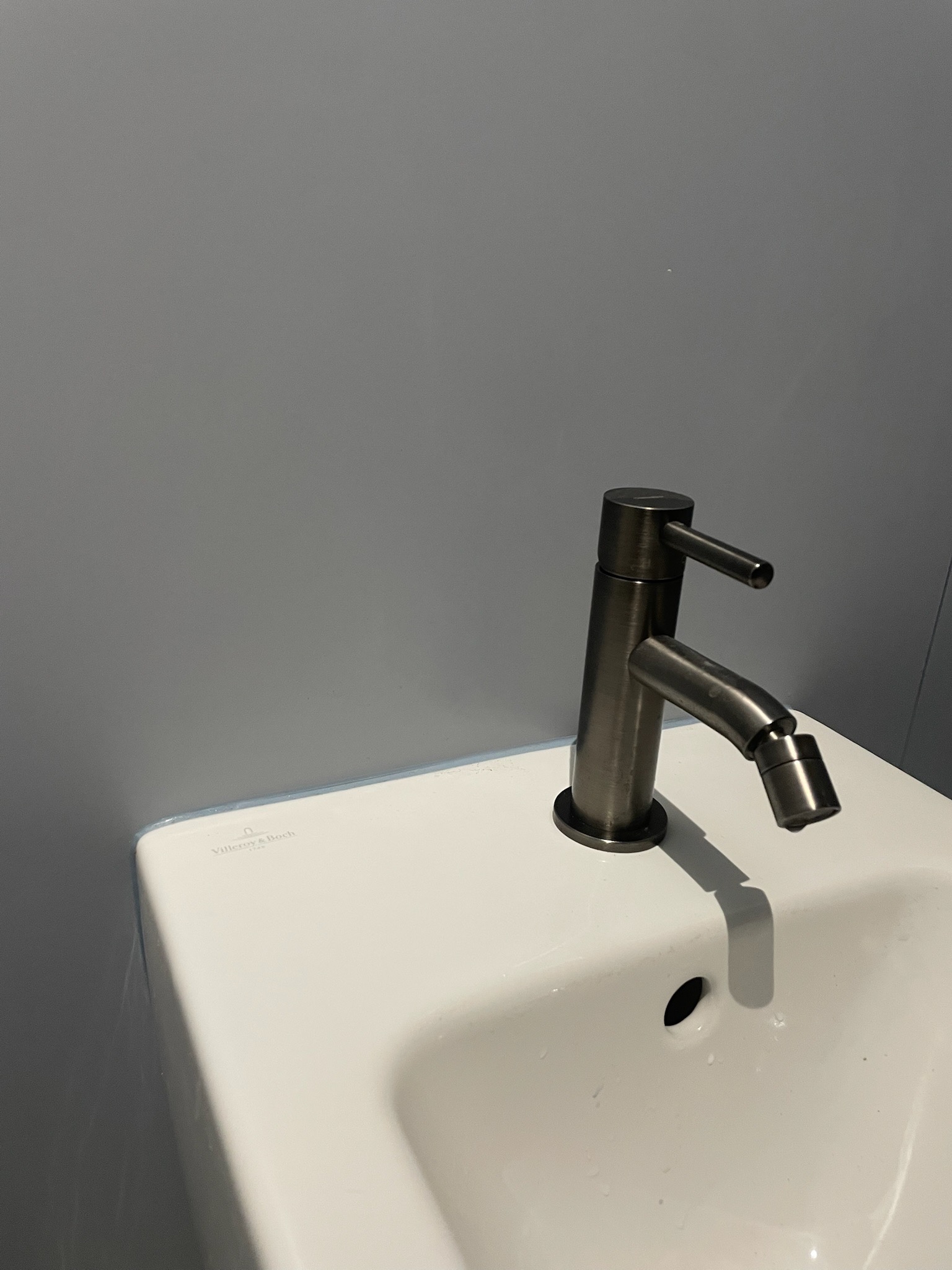 Bateria umywalkowa Villeroy & Boch w odcieniach szarości na białej umywalce, minimalistyczny design, szara ściana w tle. Ujęcie z bliska.