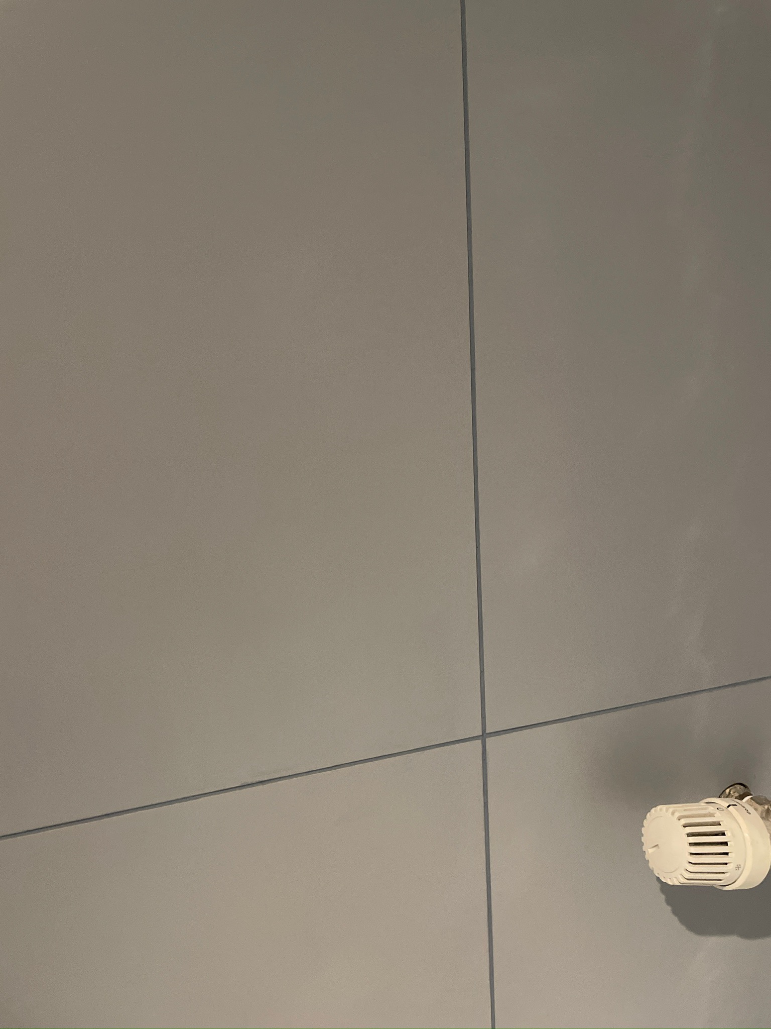 Szare płytki ścienne z termostatem w rogu. Minimalistyczny design łazienki po remoncie, z widocznymi fugami i elementami instalacji grzewczej.