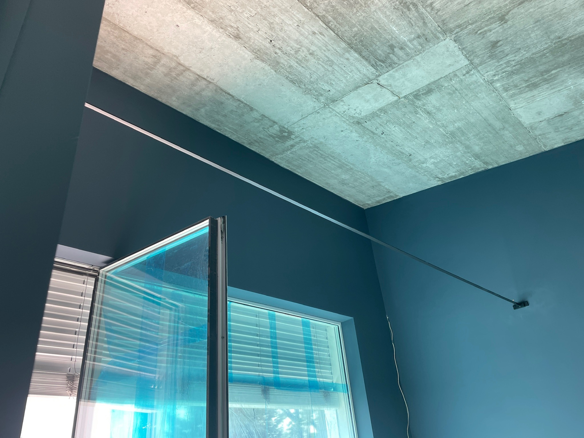 Niebieskie wnętrze z betonowym sufitem, oknem z żaluzjami i folią ochronną. Widoczna listwa oświetleniowa. Ujęcie z dołu, podkreślające surowość wykończenia.