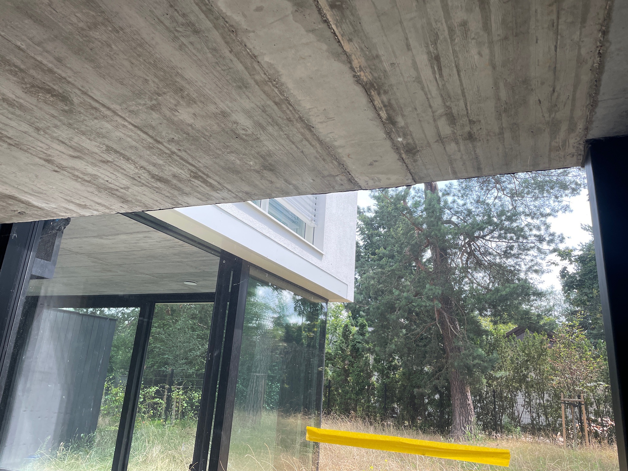 Surowy betonowy strop z odciskiem desek, widok z wnętrza przez szklaną ścianę na zieleń. Minimalistyczny design, naturalne światło.