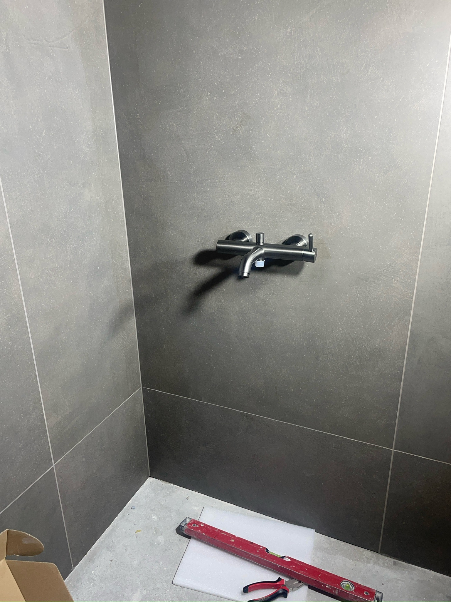 Szara, matowa łazienka w trakcie wykańczania. Widoczna bateria, płytki i poziomica z narzędziami na podłodze. Minimalistyczny design, surowy klimat.
