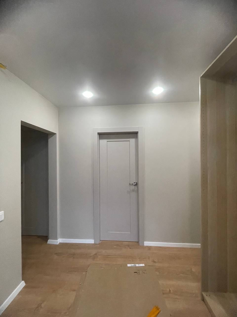 Jasne wnętrze w trakcie wykańczania: drewniana podłoga zabezpieczona kartonem, szara ściana z drzwiami i listwami przypodłogowymi. Widoczne oświetlenie sufitowe.