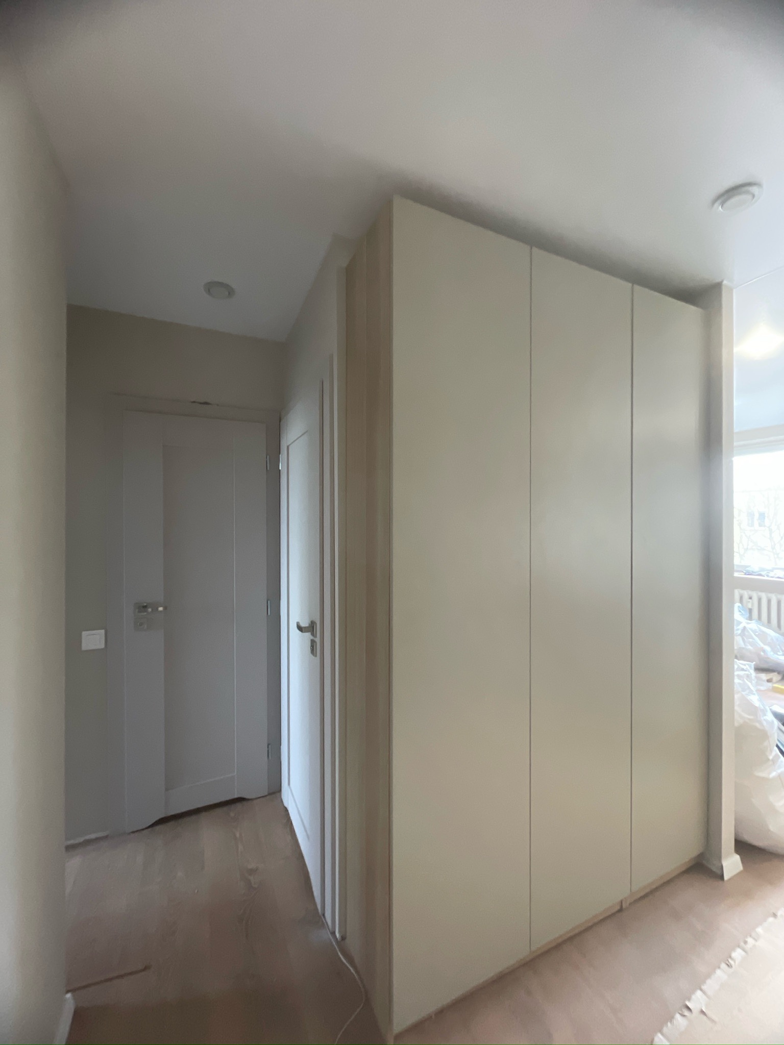 Jasne, minimalistyczne wnętrze z zabudowaną szafą wnękową i dwiema parami drzwi. Parkiet na podłodze, neutralna kolorystyka ścian i mebli. Widok na okno w tle.