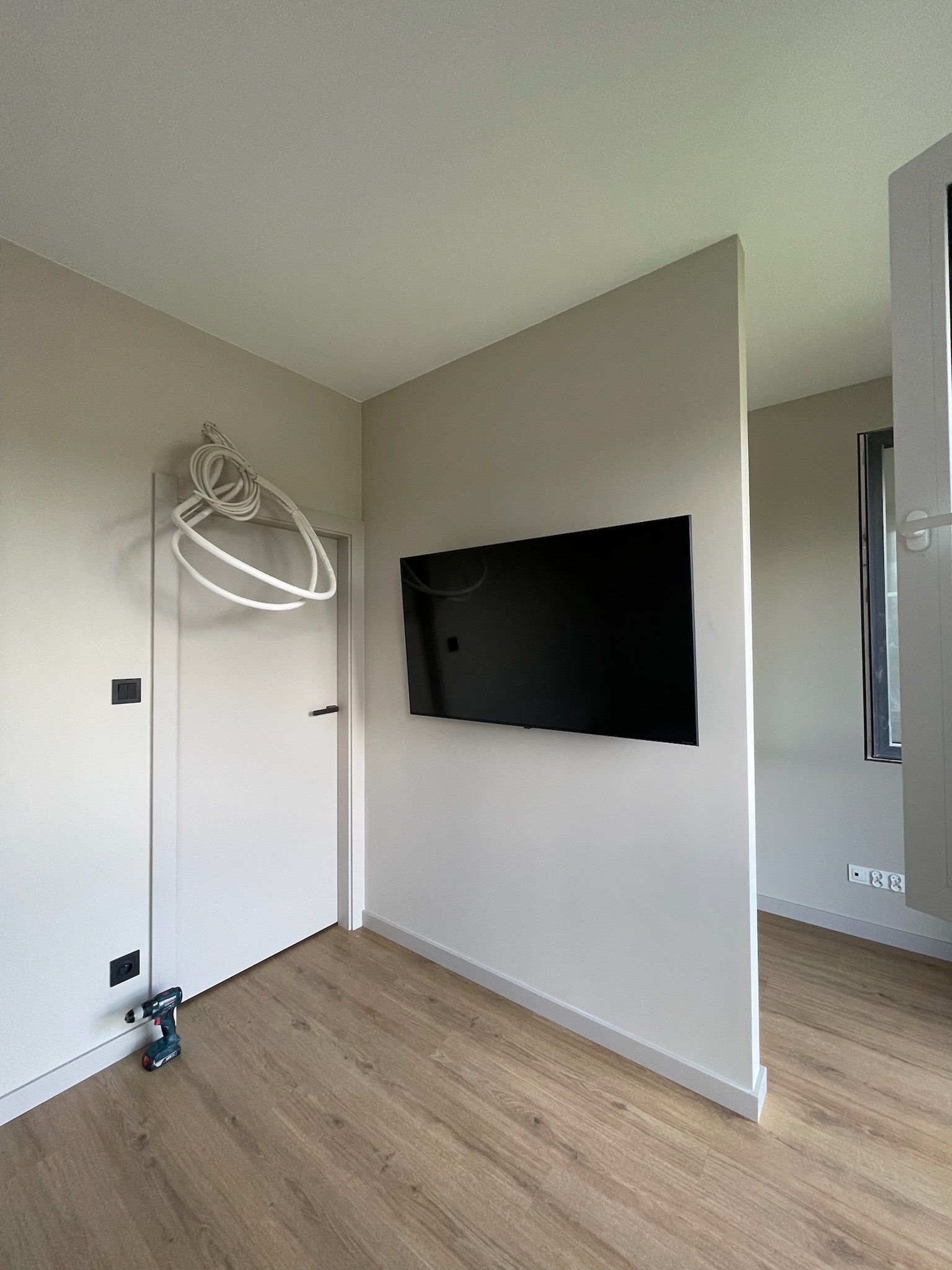 Minimalistyczne wnętrze po montażu TV: ściana działowa, telewizor, kabel zwisający z drzwi, wiertarka przy ścianie, jasna podłoga i listwy przypodłogowe.