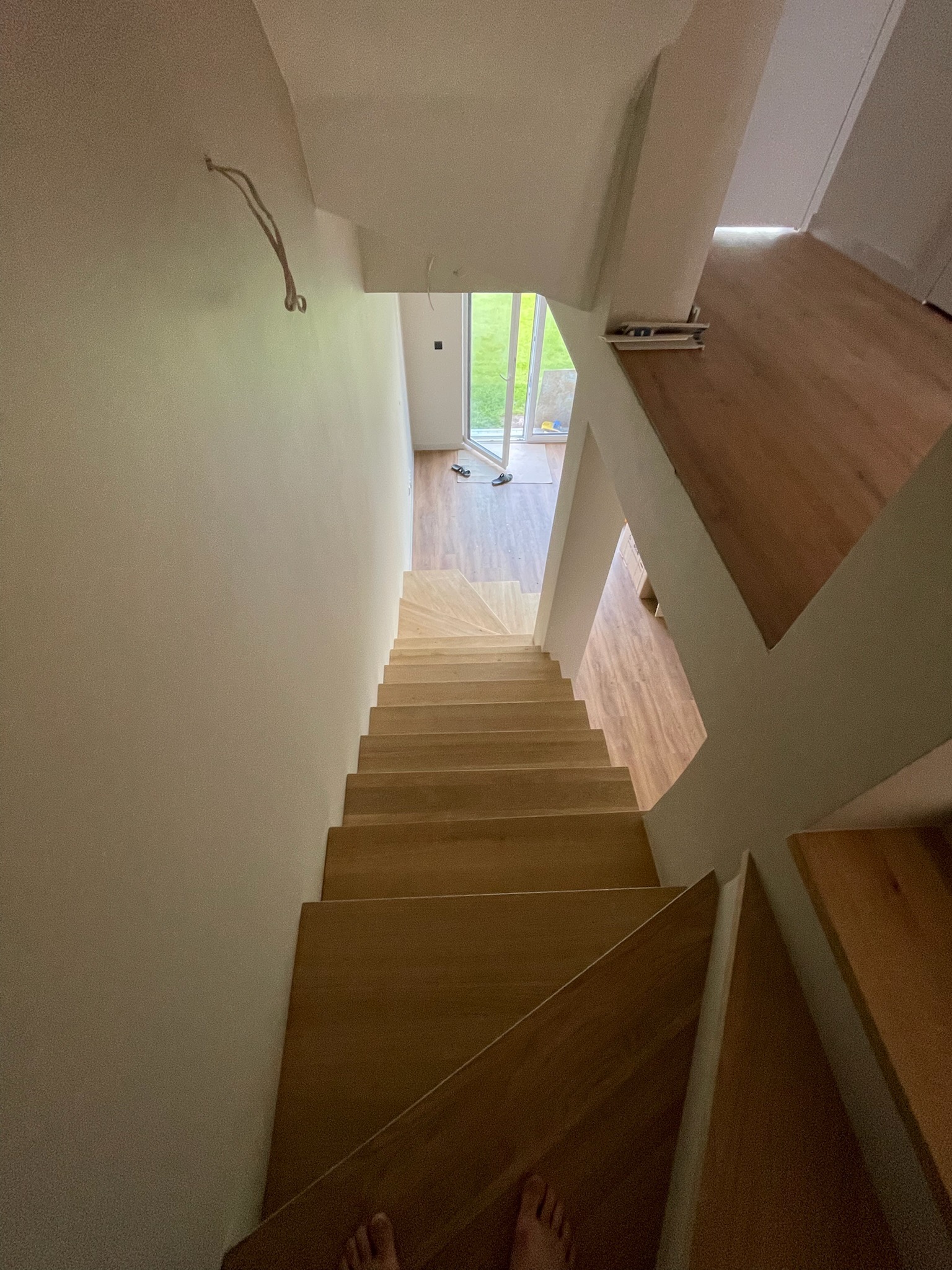 Drewniane schody w jasnym wnętrzu, widok z góry. Minimalistyczny design, białe ściany, naturalne światło wpadające przez okno. Częściowo widoczne stopy na dole kadru.