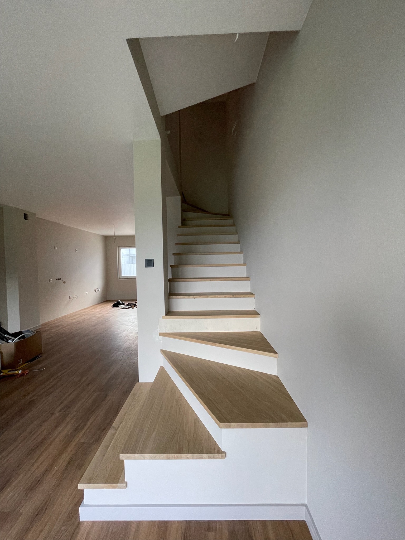 Drewniane schody z białymi podstopnicami w jasnym wnętrzu domu. Widoczny fragment salonu z drewnianą podłogą i ścianami w trakcie wykończenia.